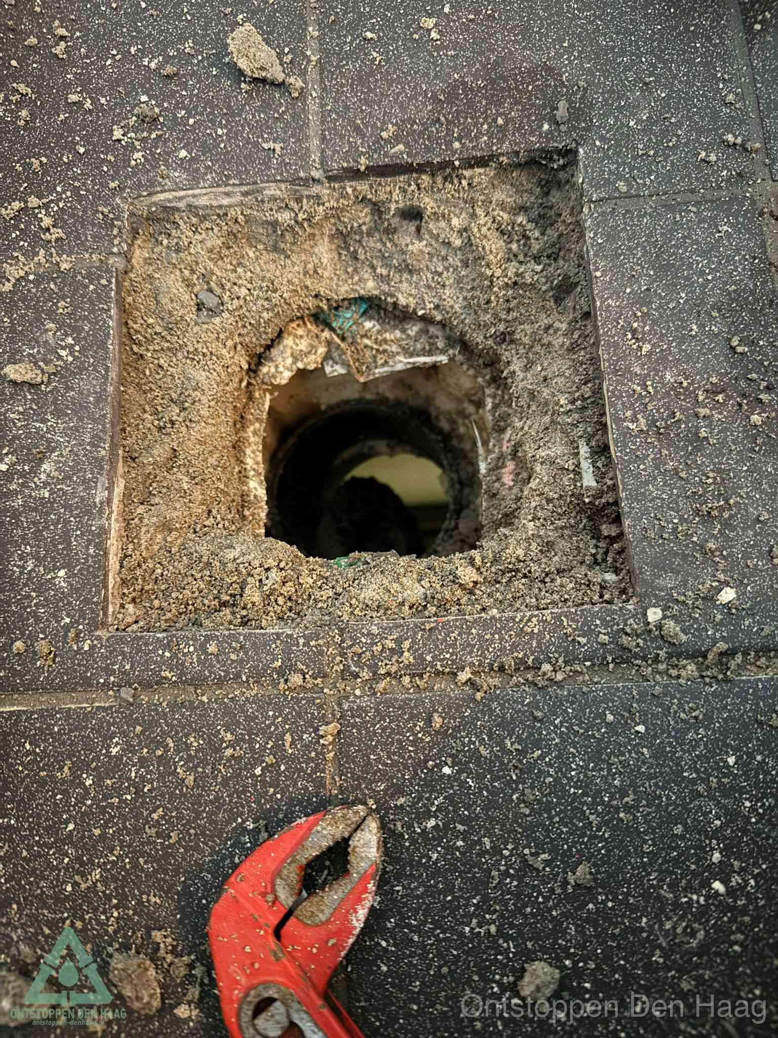 Een geopend rioolputje in een betontegelvloer met losgemaakt rooster en een zichtbare ronde afvoerbuis, het achterhalen waar de afvoerverstopping zit in Den Haag