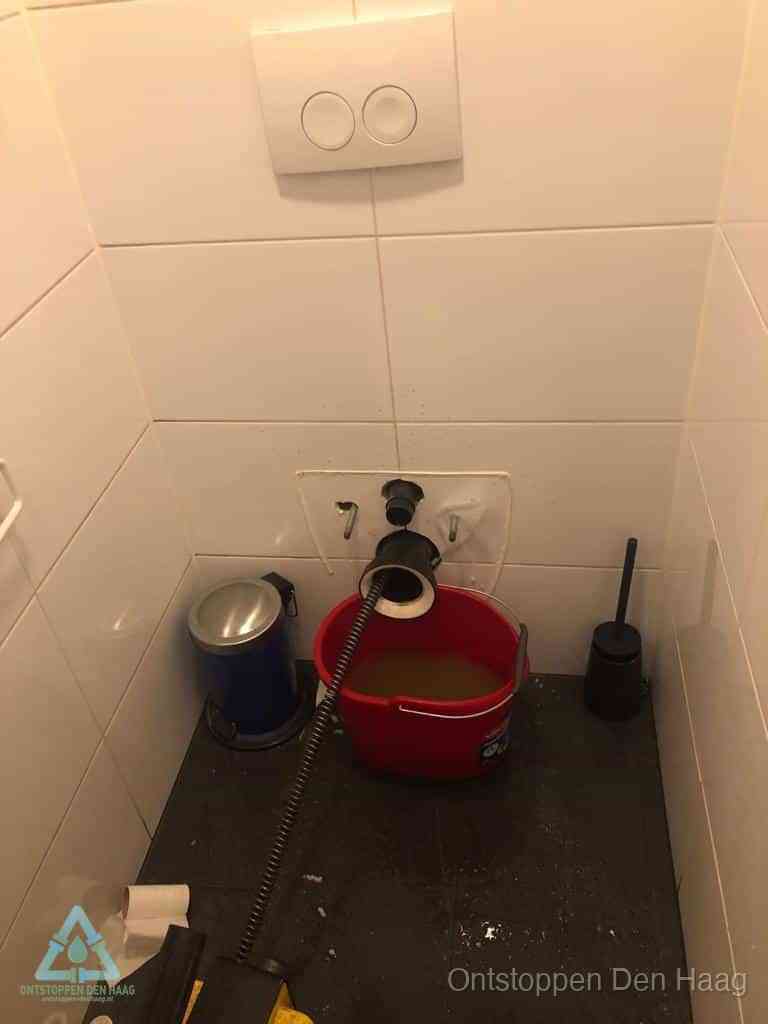 Een ontstopte wand-wc met verwijderde toiletpot waar een ontstoppingsveer in de wanduitgang verdwijnt, het achterhalen waar de verstopping zit in Den Haag