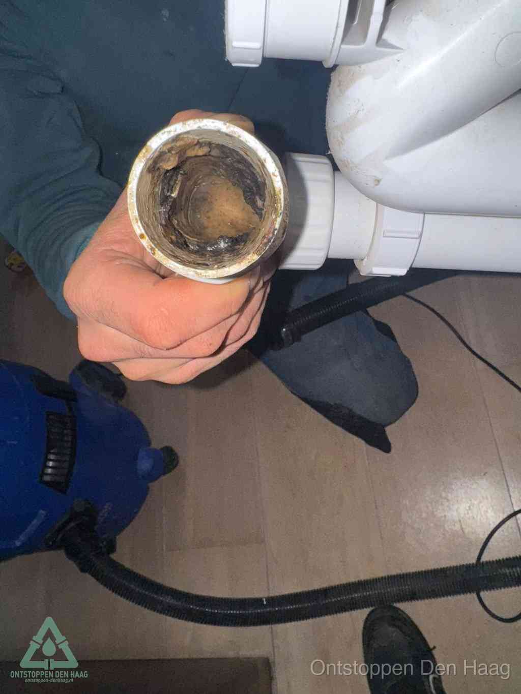 Een hand houdt een losgekoppeld PVC-afvoerbuisstuk vast dat van binnen volledig is dichtgeslibd met vet, vuil en aanslag, de meest voorkomende oorzaak van een verstopte afvoer in Den Haag