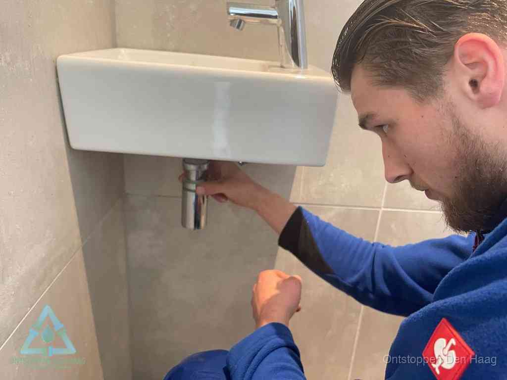 Een monteur draait met de hand de zwanenhalssifon onder een fonteinwasbak in de toiletruimte los om de bron van rioollucht in Den Haag te vinden