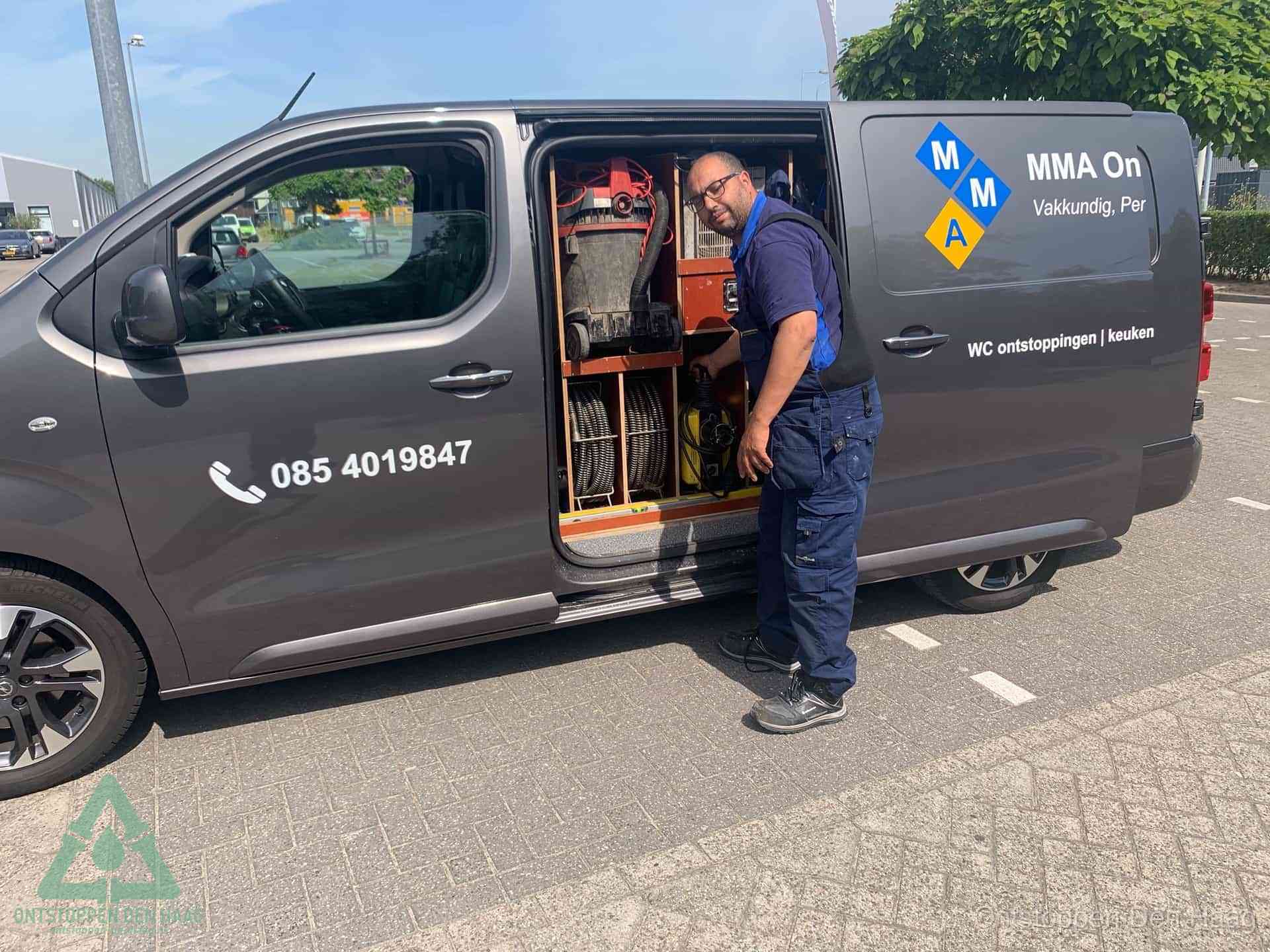 Een monteur bij de open schuifdeur van zijn bedrijfsbus op een parkeerplaats, klaar om de oorzaak van terugkerende afvoerverstoppingen aan te pakken in Den Haag
