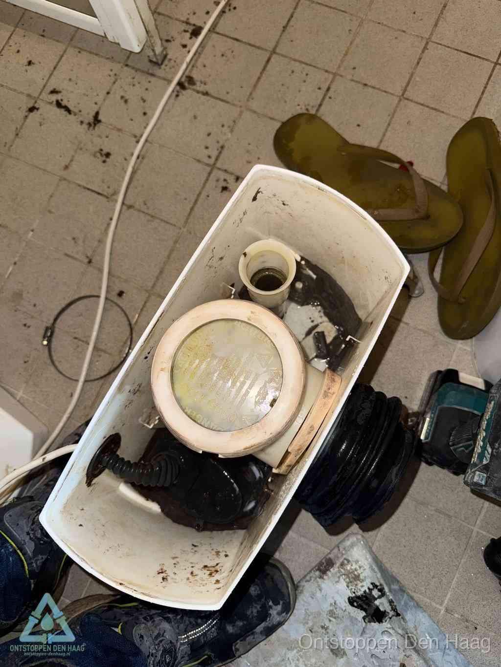 Een gedemonteerde toiletpot op de vloer met ontstoppingsveer en gereedschap ernaast, het verwijderen van een vastzittend voorwerp uit de wc in Den Haag