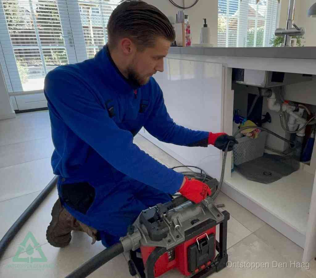 Een monteur met rode handschoenen knielt in een keuken en voert een elektrische ontstoppingsveer in tijdens het professioneel afvoer ontstoppen in Den Haag