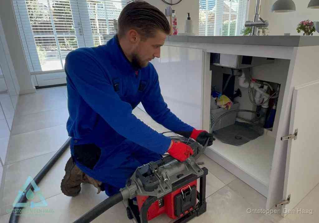 Een monteur voert een professionele elektrische ontstoppingsmachine in de afvoer onder de spoelbak tijdens het gootsteen ontstoppen in Den Haag
